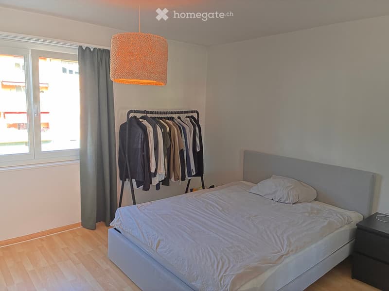 Cozy bedroom with a double bed, nightstand, hanging clothes, and a large window with a curtain.