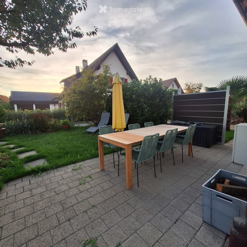 Garden area with a wooden table, green chairs, umbrella, and surrounding greenery.