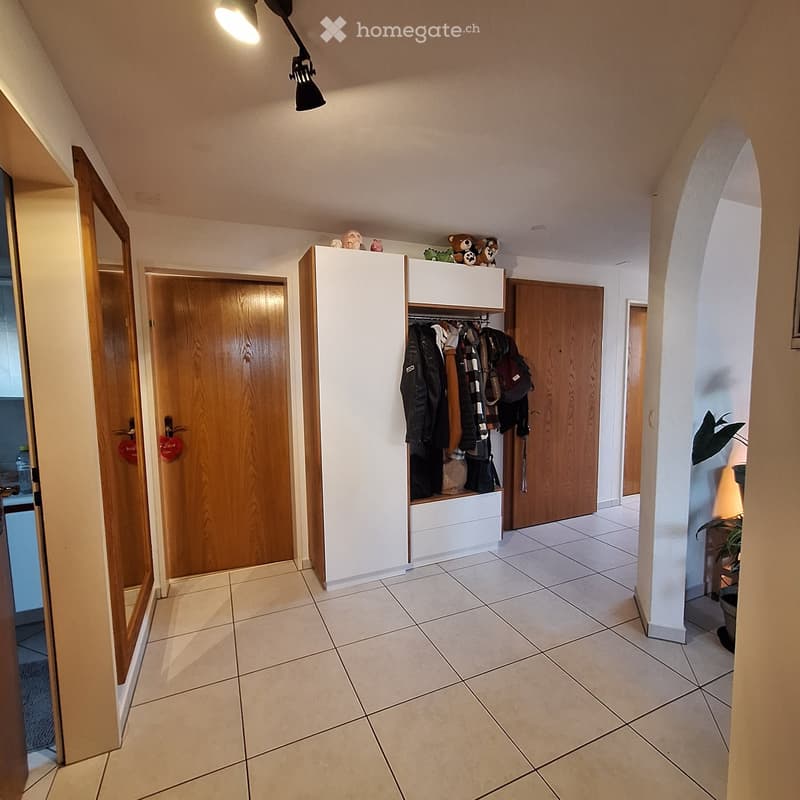 Hallway with tiled floor, white walls, wooden doors, and a white coat rack with hanging items and stuffed toys on top.