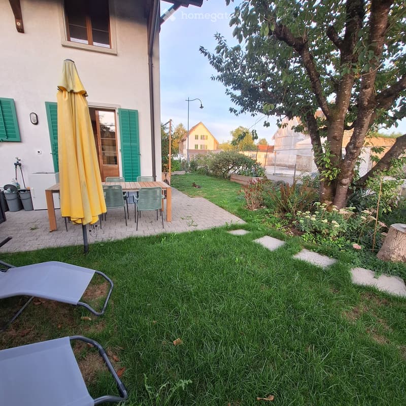 Garden with green lawn, patio area with table and chairs, umbrella, lounge chairs, and a tree.