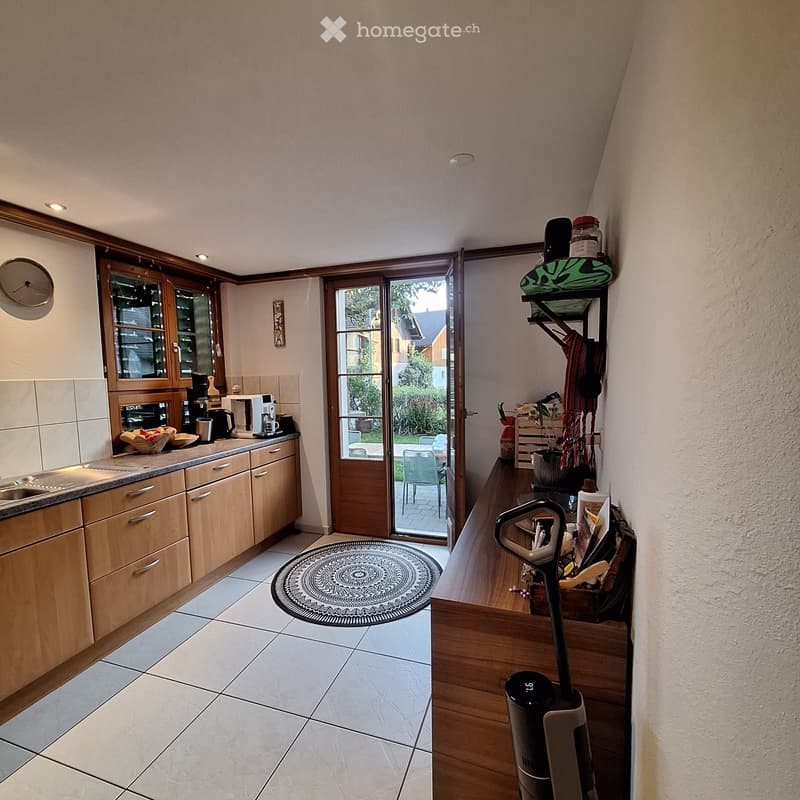 Bright kitchen with wooden cabinets, tiled floor, and a round patterned rug. A sliding glass door leads to a patio with outdoor furniture.