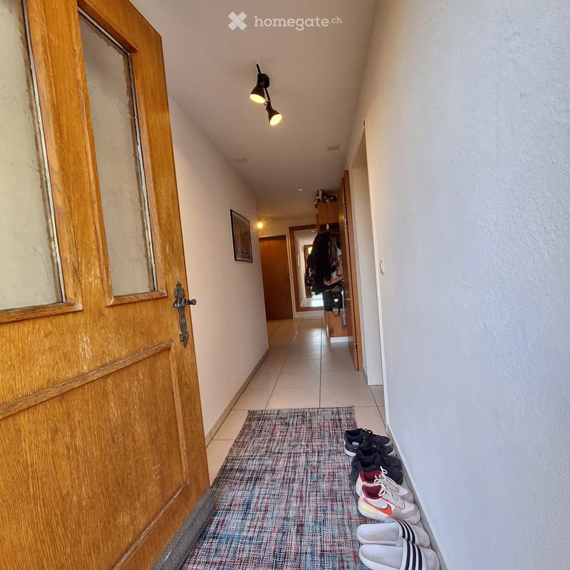 A narrow hallway with tiled flooring, a patterned rug, and several pairs of shoes placed against the wall. A wooden door is visible on the left, and a small wall-mounted light fixture illuminates the space.