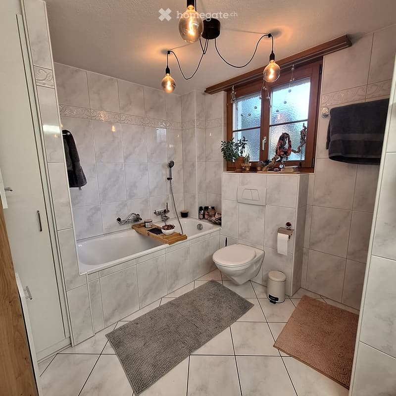 Modern bathroom with a bathtub, toilet, sink, and mirror. Features tiled walls, wooden window frame, and decorative lighting.