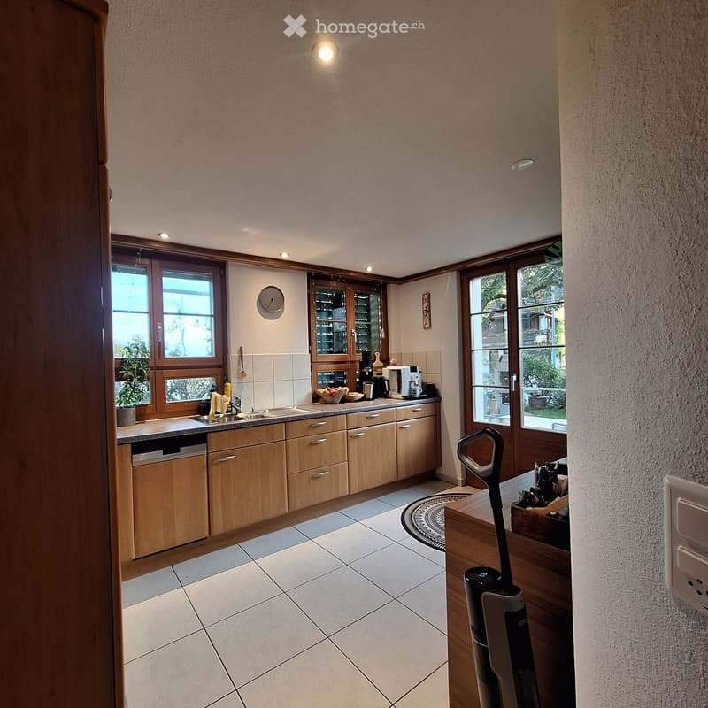 Bright kitchen with wooden cabinets, tiled floor, and large windows. Includes a sink, dishwasher, coffee maker, microwave, and a vacuum cleaner in the foreground.