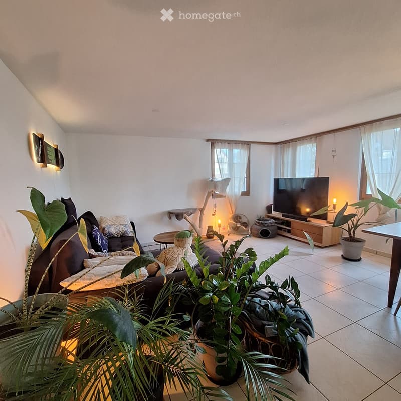 A cozy living room with a dark brown sofa, a coffee table, a television on a wooden stand, a fan, a cat tree, and several potted plants. The room has large windows with white curtains and a slanted ceiling.