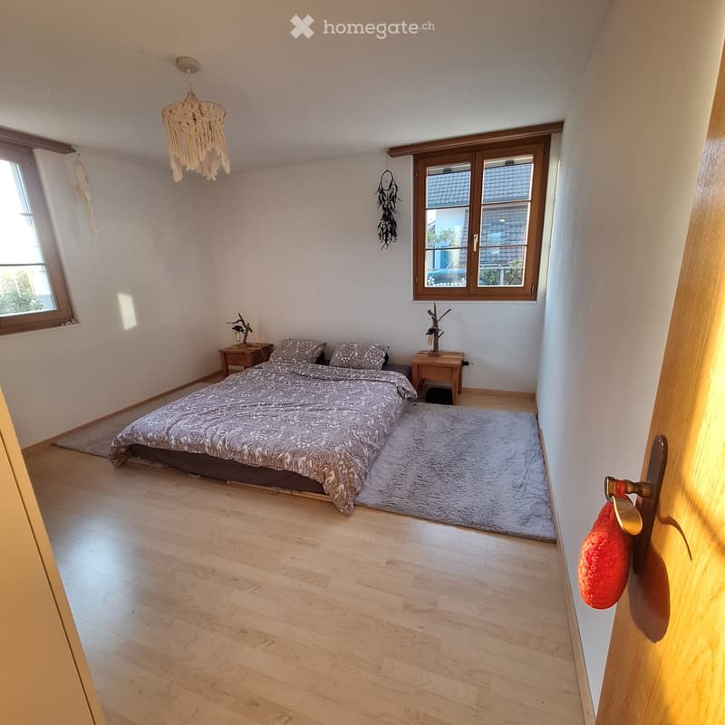Cozy bedroom with wooden flooring, large windows, a bed with patterned bedding, nightstands, a wicker chandelier, and a dreamcatcher on the wall.