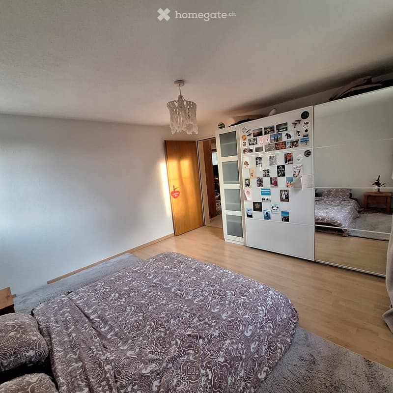 A modern bedroom with a large bed covered in patterned bedding, a full-length mirror, a wardrobe with photos and decorations, and a nightstand with a lamp.