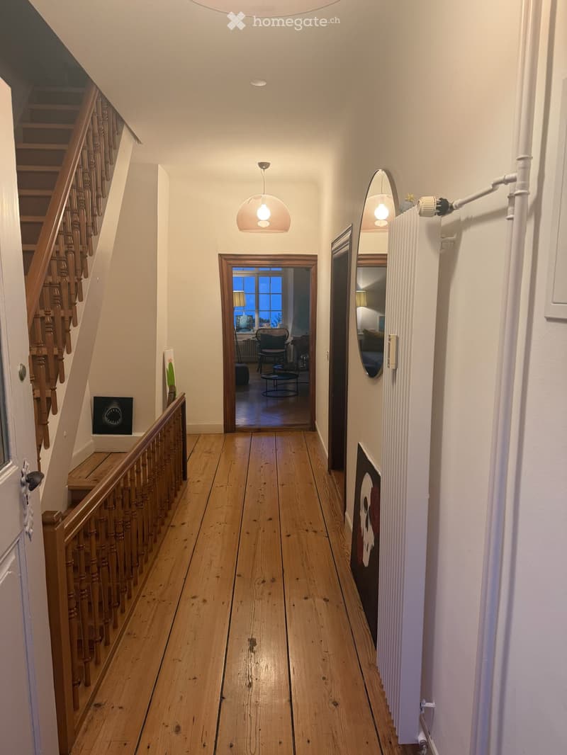 Wooden floor hallway with staircase on left, pendant light, mirror, radiator, and open doorway leading to another room with visible furniture.