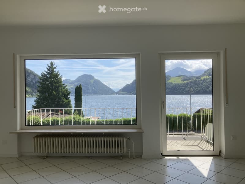 Empty room with large windows offering scenic views of a lake and mountains, tiled floor, radiator, and sliding glass door leading to a balcony.