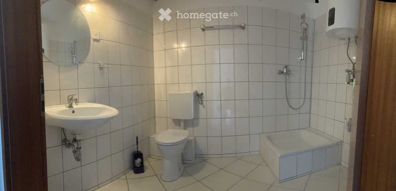 A small bathroom with white tiled walls and floor, featuring a sink, toilet, bathtub, and a mirror above the sink. A showerhead is mounted on the wall, and a handrail is installed for safety.