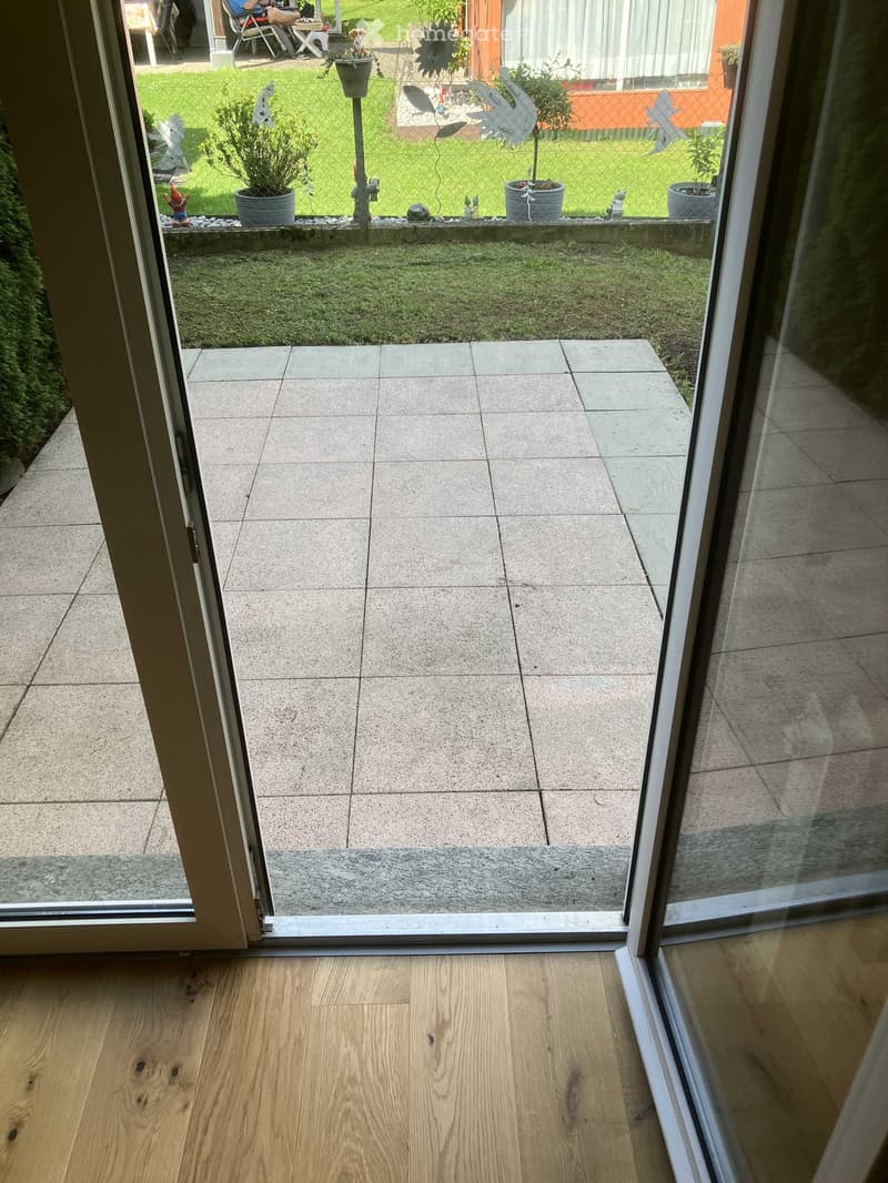 View from inside a house through a large sliding glass door onto a tiled balcony area leading to a small garden with grass, potted plants, and decorative items.