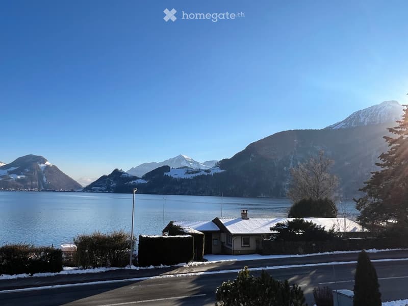 A small house with a snow-covered roof sits near a road, surrounded by trimmed hedges and trees, with a serene lake and snow-capped mountains in the background under a clear blue sky.