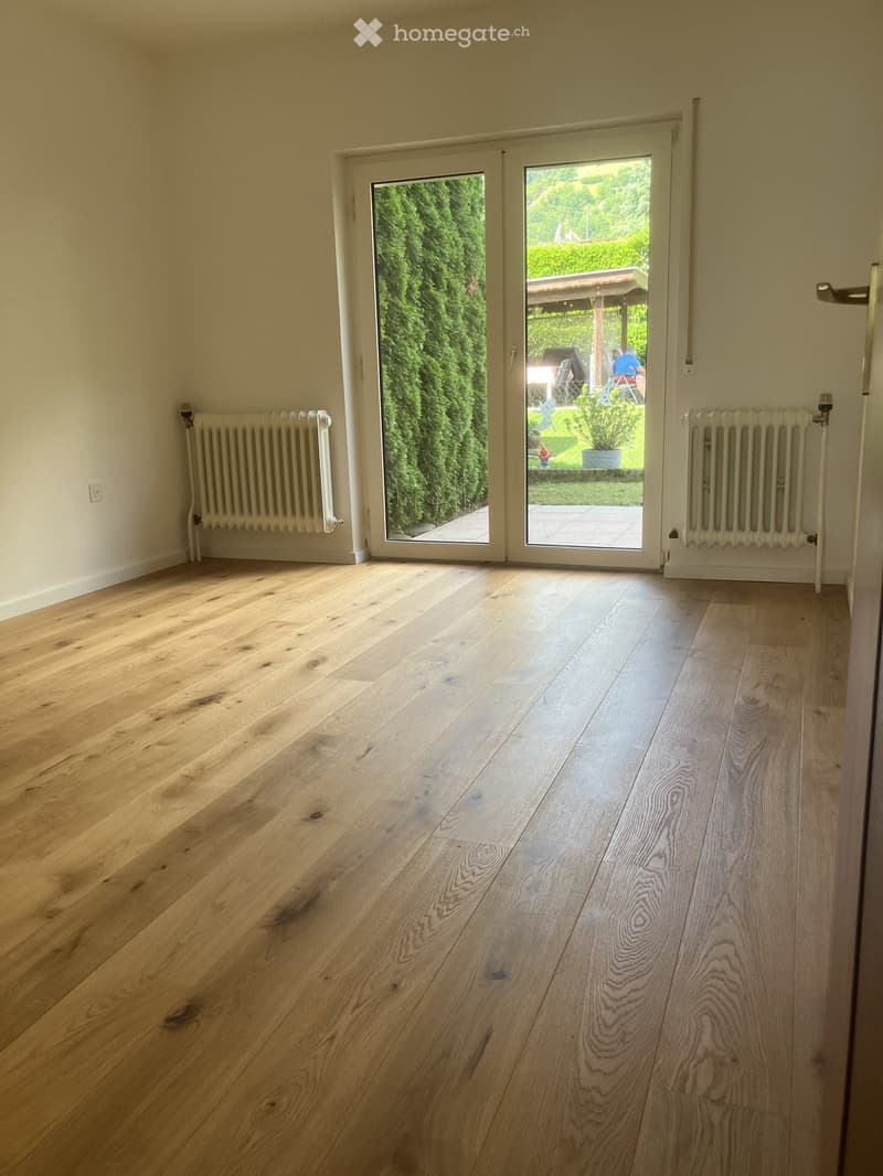 Empty room with light wooden flooring, white walls, radiators on both sides, and large glass sliding doors leading to a garden area.