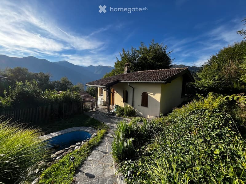 A small, single-story house with a tiled roof and light yellow walls, surrounded by a well-maintained garden with a swimming pool and lush greenery, offering a scenic mountain view in the background.