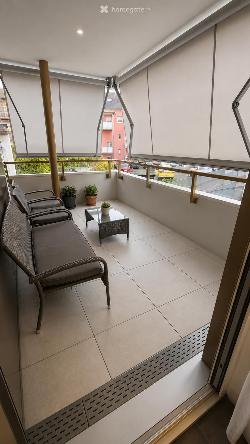 Balcony with tiled floor, white walls, and large windows with blinds. Features a wicker sofa, small glass table, and potted plants.