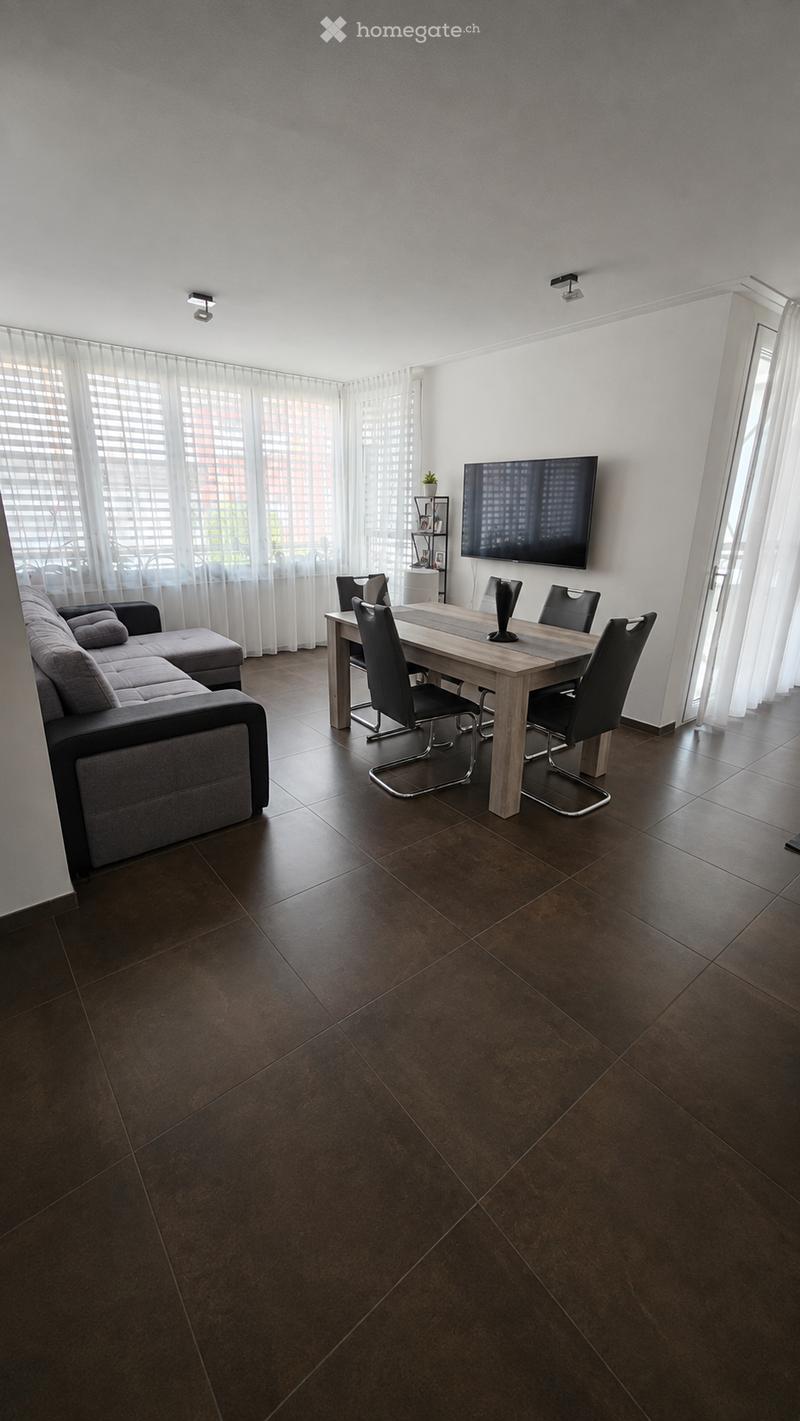 Modern living and dining area with dark brown tiled flooring, large windows with sheer curtains, a grey sectional sofa with an ottoman, a wooden dining table with four black chairs, a flat-screen TV mounted on the wall, and a small plant on a shelf.