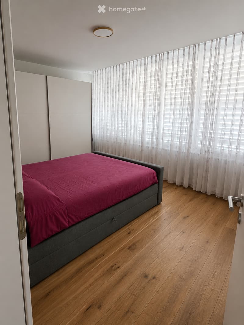 Modern bedroom with wooden flooring, a grey bed with a pink duvet, white curtains, and a built-in wardrobe.