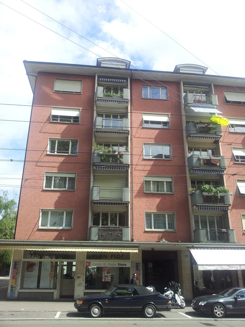 A multi-story residential building with pink facade, multiple windows, balconies with plants, and storefronts at the ground level including a Canon poster store and a fashion boutique.