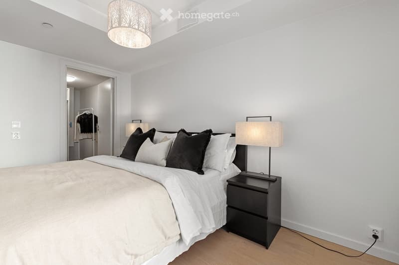 Modern bedroom with a large bed, white bedding, black pillows, a nightstand with a lamp, and a decorative pendant light hanging from the ceiling.