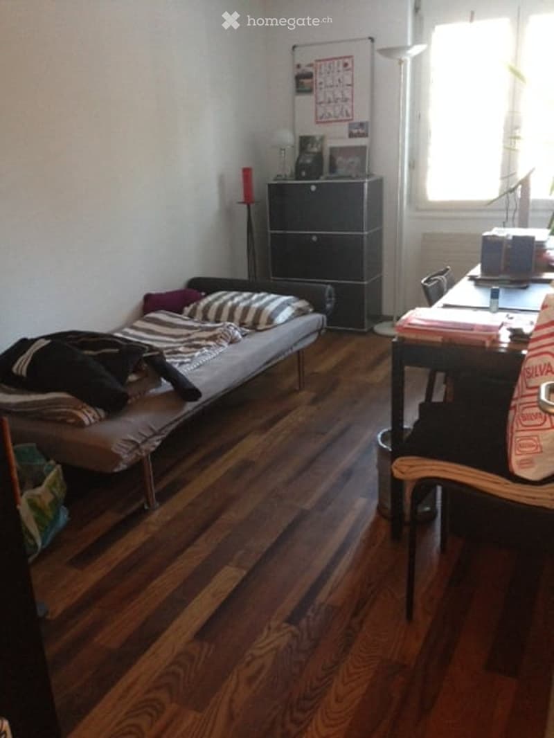 A small, furnished room with a sofa bed, a chair, a table, a cabinet, and a lamp. The room has wooden flooring and white walls.