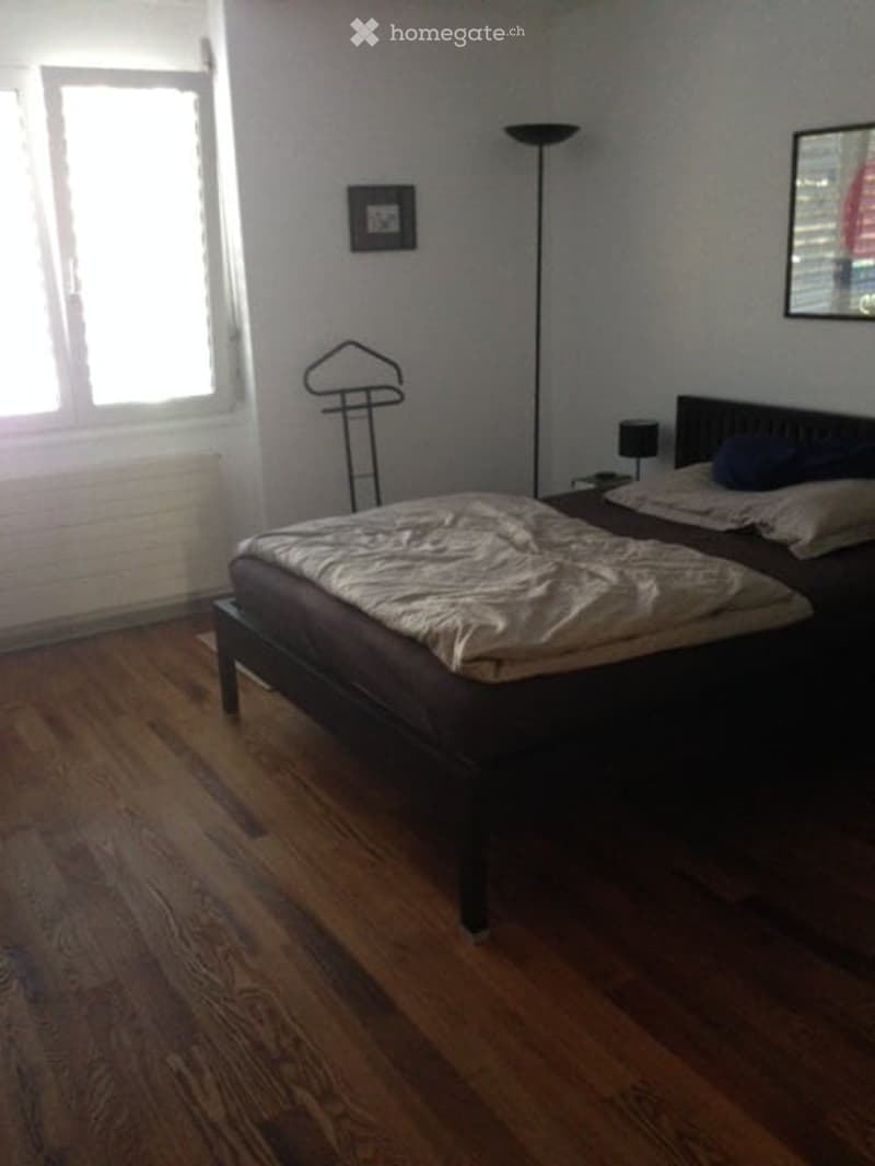Bedroom with wooden flooring, a dark wooden bed with white bedding, a nightstand, a floor lamp, a mirror, and a coat rack.