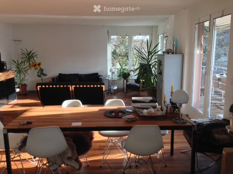 Bright, open-plan living and dining area with wooden flooring, large windows, and modern furniture including a dining table, chairs, sofa, and refrigerator.