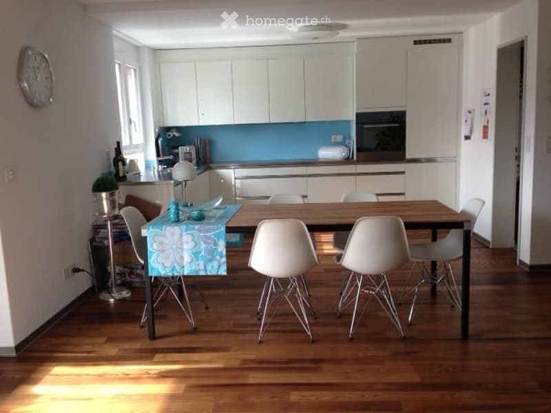 Modern kitchen with white cabinets, a wooden dining table surrounded by white chairs, and a blue accent wall. A small plant and a bottle are placed on the counter.