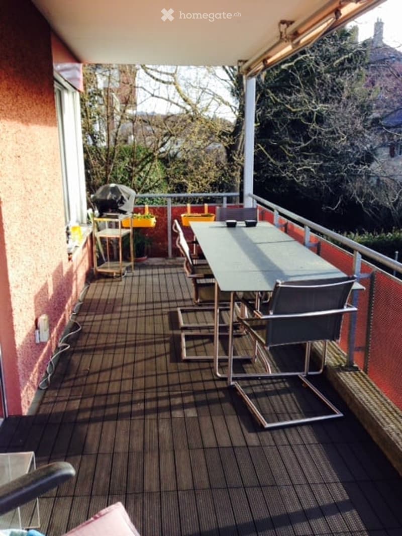Balcony with wooden decking, metal railing, and outdoor furniture including a table and chairs, overlooking a garden and trees.