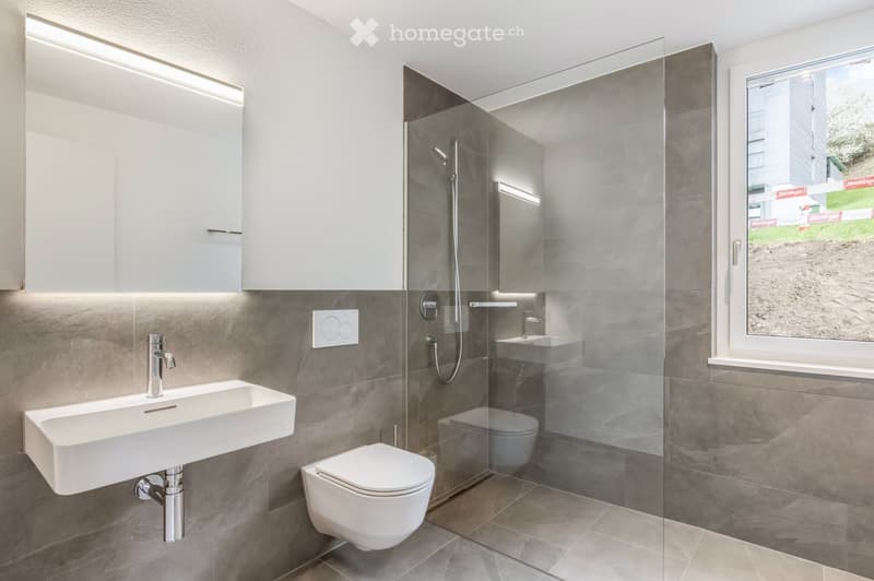Modern bathroom with a white sink, toilet, and glass-enclosed shower. Features grey tiles, built-in cabinets, and a large window offering a view of the outside.