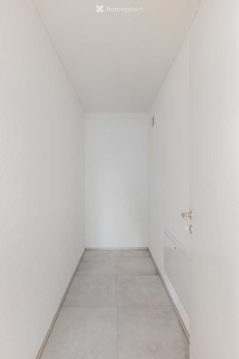 A clean, minimalist hallway with white walls, a grey tiled floor, and a small built-in cabinet on the right side.