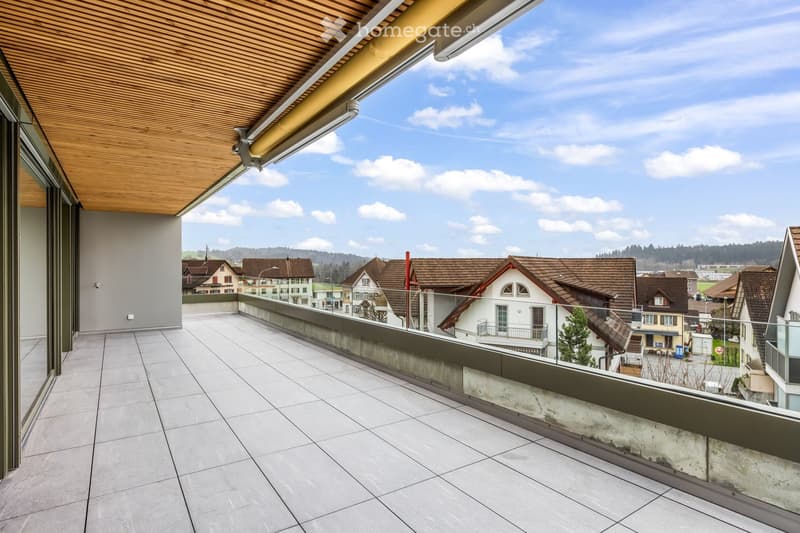 Balcony with tiled floor, wooden ceiling, glass railing, overlooking a residential area with houses and a clear sky.