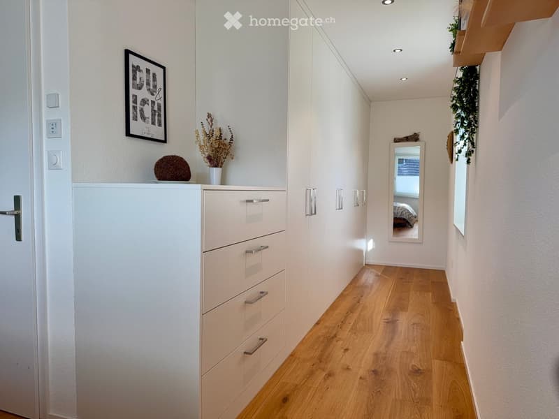 Modern hallway with light wooden flooring, white walls, built-in storage units, and decorative items including a framed picture, a vase with dried flowers, and a small round decorative object.