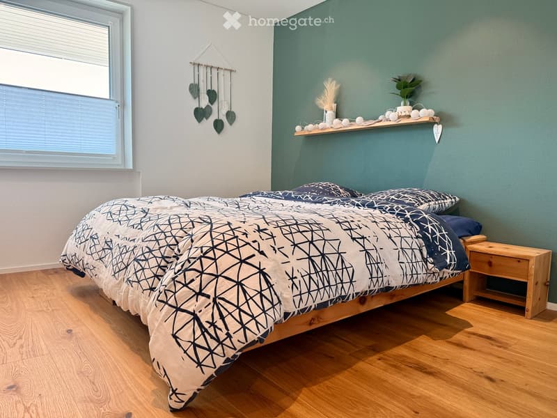 Cozy bedroom with a wooden bed, geometric-patterned duvet, nightstand, and decorative wall hanging. Green accent wall and wooden flooring.