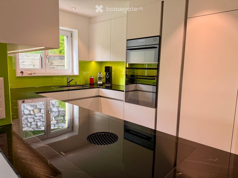 Modern kitchen with white cabinets, green backsplash, black countertops, and stainless steel appliances including an oven and coffee maker.