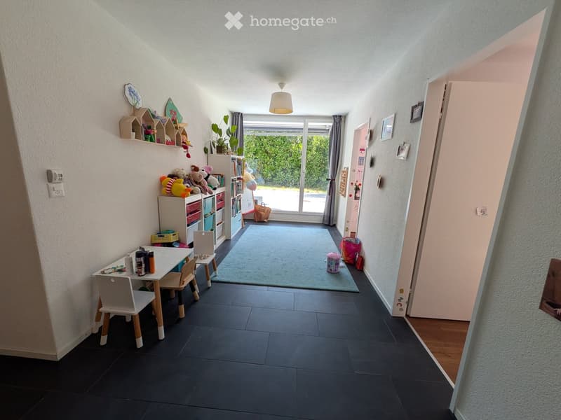 Brightly lit room with dark tiled floor, white walls, and large window with view to the garden. Contains a small table with chairs, shelves with toys, and a rug in the center.