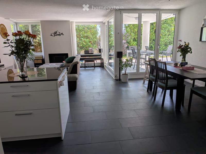 Bright, open living and dining area with large windows and sliding glass doors leading to a patio. Features a sofa, coffee table, dining table with chairs, kitchen cabinetry, and decorative plants.