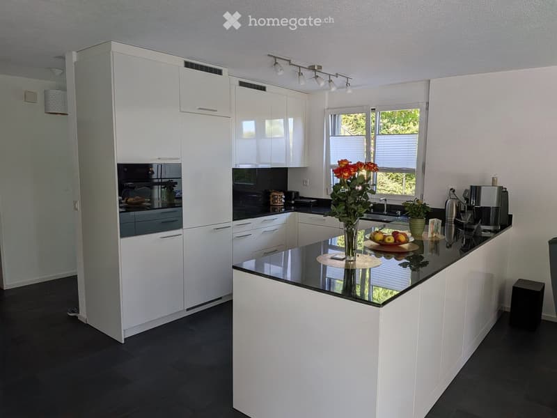Modern kitchen with white cabinets, black countertops, and stainless steel appliances. Features a central island with a vase of flowers and a bowl of fruit, along with a coffee maker and a potted plant on the counter.