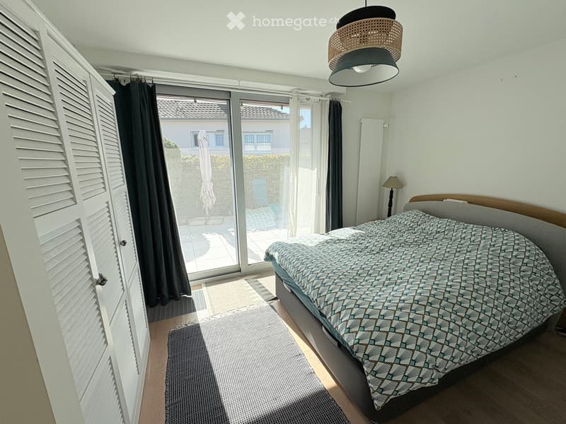 Bedroom with a double bed, patterned bedspread, a small rug, and a hanging lamp. Large sliding glass doors lead to a patio with outdoor furniture.
