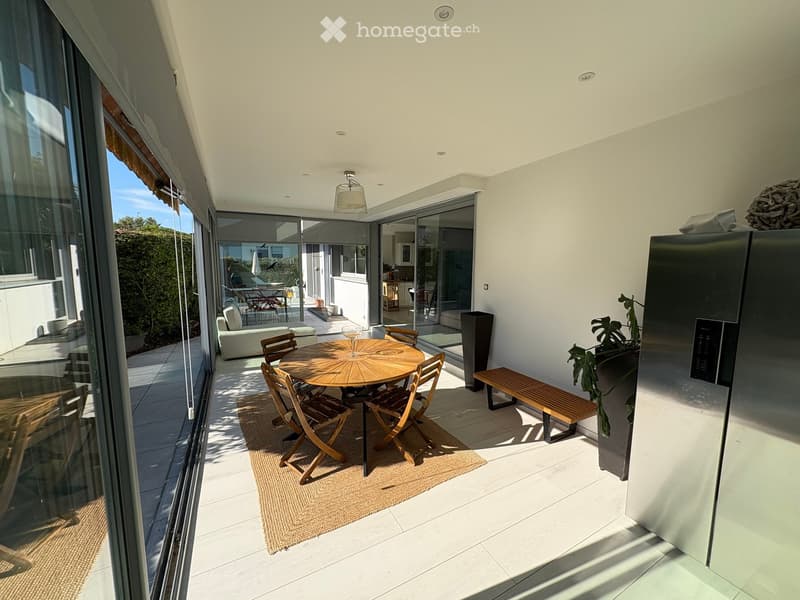 Modern outdoor terrace with a round wooden table, chairs, a bench, and a sofa visible through large glass doors. The space features a tiled floor, a hanging light fixture, and a small plant near the refrigerator.