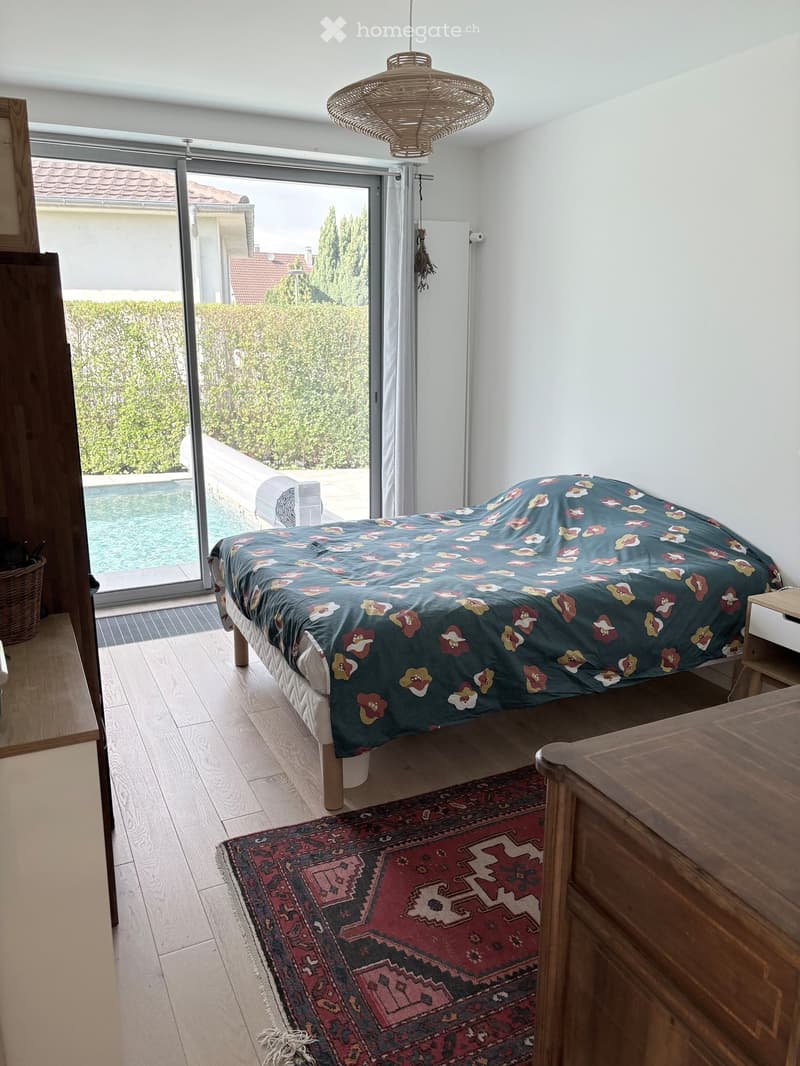 Bright bedroom with wooden flooring, a large bed with a patterned duvet, a nightstand, a wicker basket, and a colorful rug. Large sliding glass doors open to a view of a swimming pool and garden.