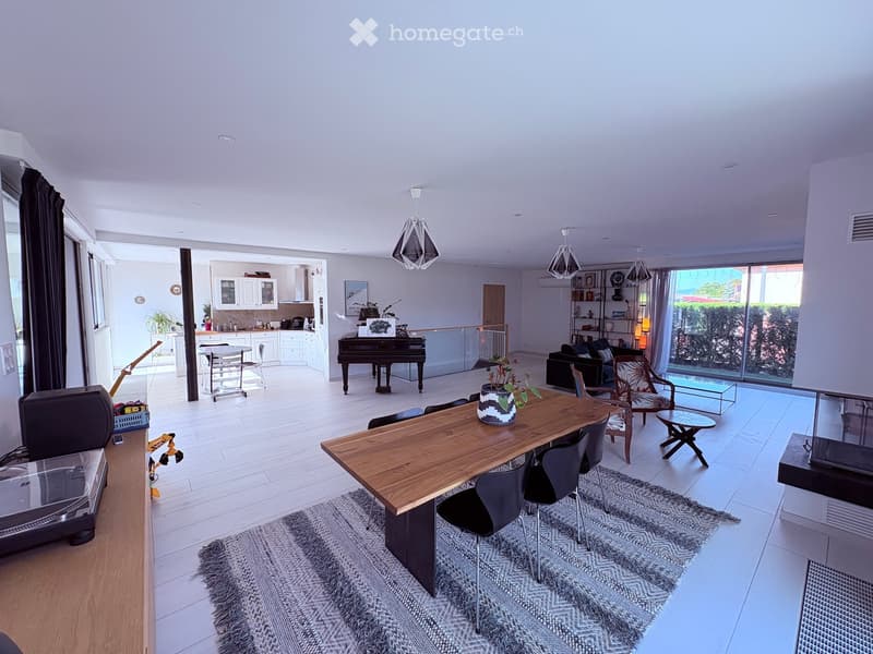 Open-plan living and dining area with a wooden dining table, black chairs, a sofa, a piano, and a modern kitchen with white cabinets and a sink.