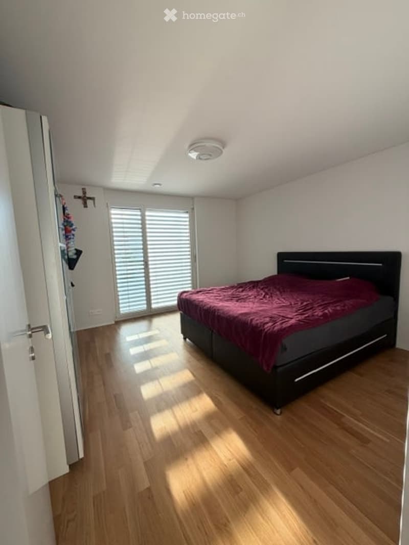 Bright bedroom with wooden flooring, large window with blinds, bed with dark frame and red bedding, and a nightstand.