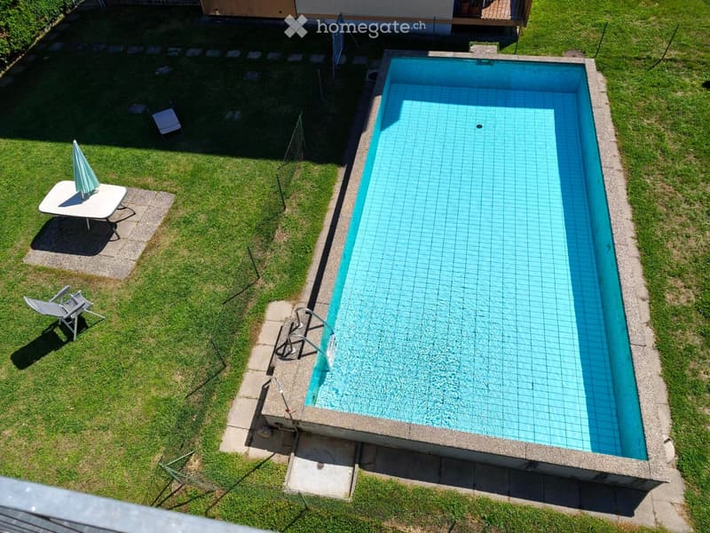 A rectangular swimming pool with clear blue water is surrounded by a concrete edge. Nearby, there is a small table with an umbrella and several lounge chairs on the grass.