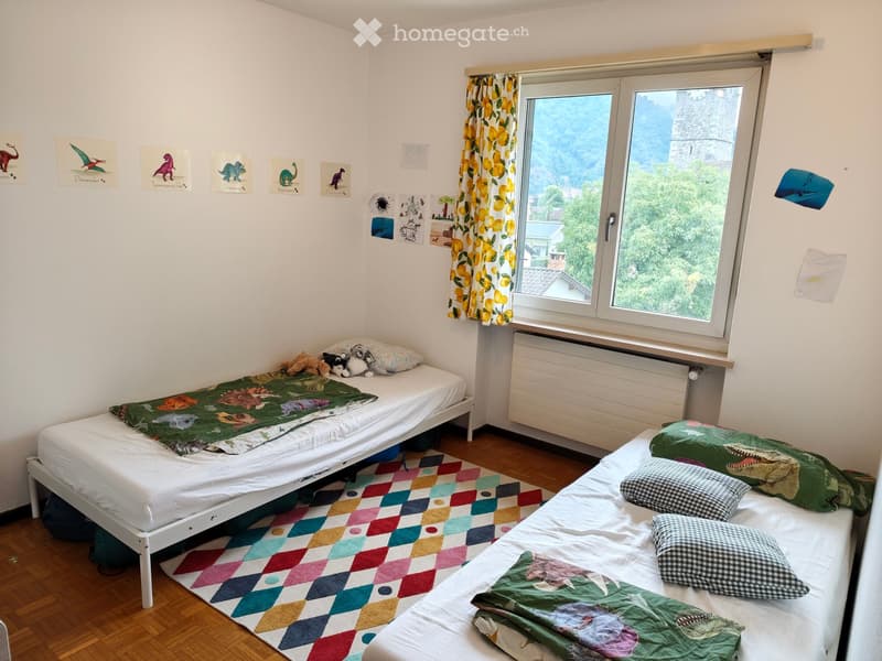 Cozy bedroom with two single beds, dinosaur-themed blankets, colorful rug, and large window with scenic view.