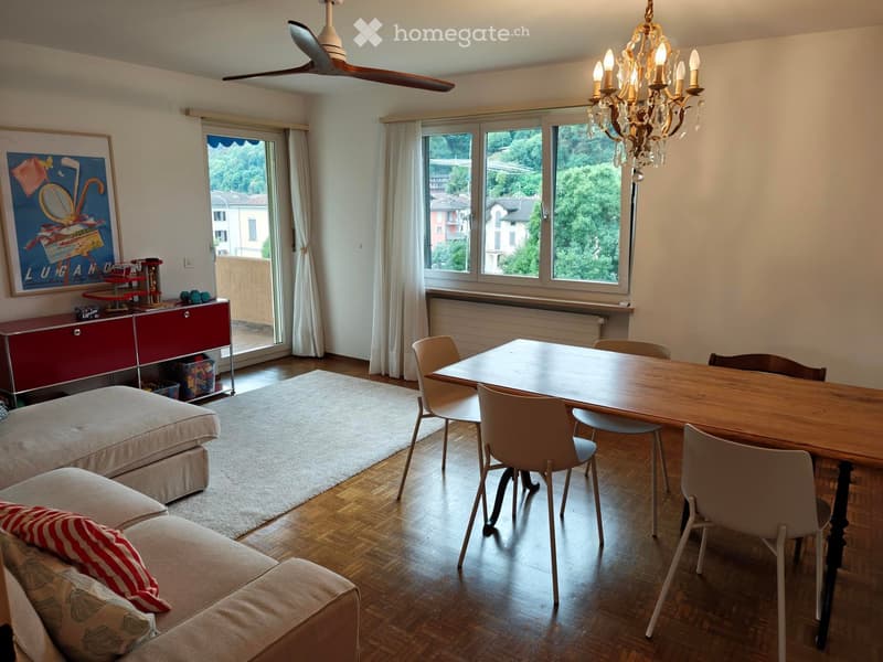 Living and dining area with a sofa, dining table, chairs, cabinet, chandelier, ceiling fan, and large windows offering a scenic view.