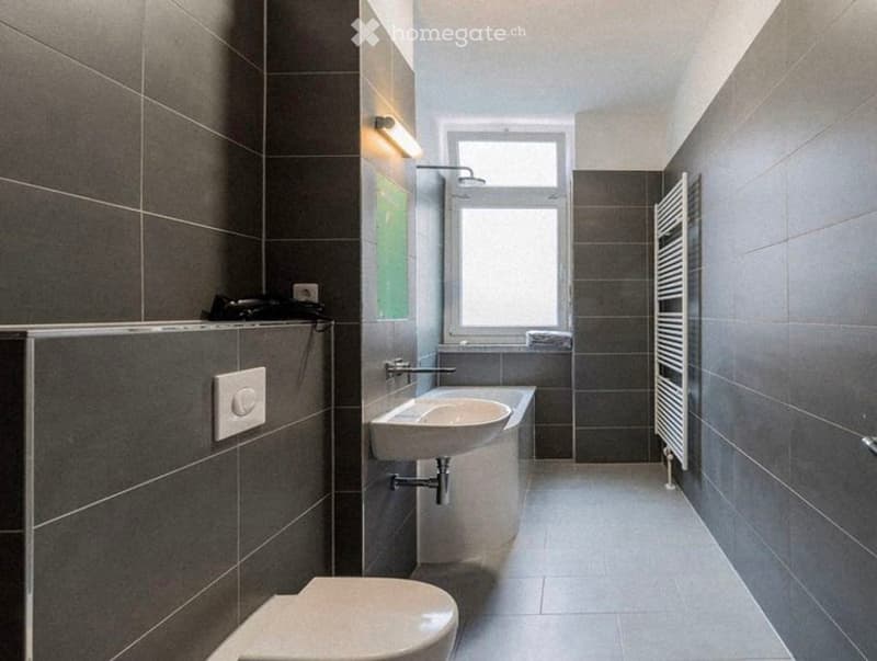 Modern bathroom with dark grey tiles, a white sink, toilet, and a window above the sink. A radiator is mounted on the wall, and a mirror is positioned above the sink.