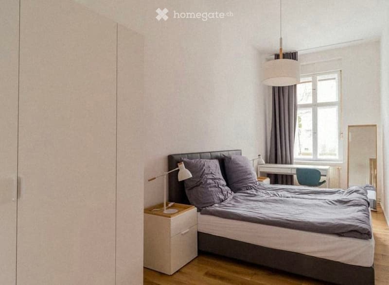 A modern, minimalist bedroom with a large bed, nightstand, desk, and chair. The room features light-colored walls, wooden flooring, and a large window with curtains.