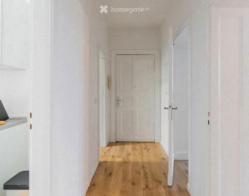 A narrow hallway with light wooden flooring and white walls, featuring a closed door at the end and an open door to the left leading to another room.