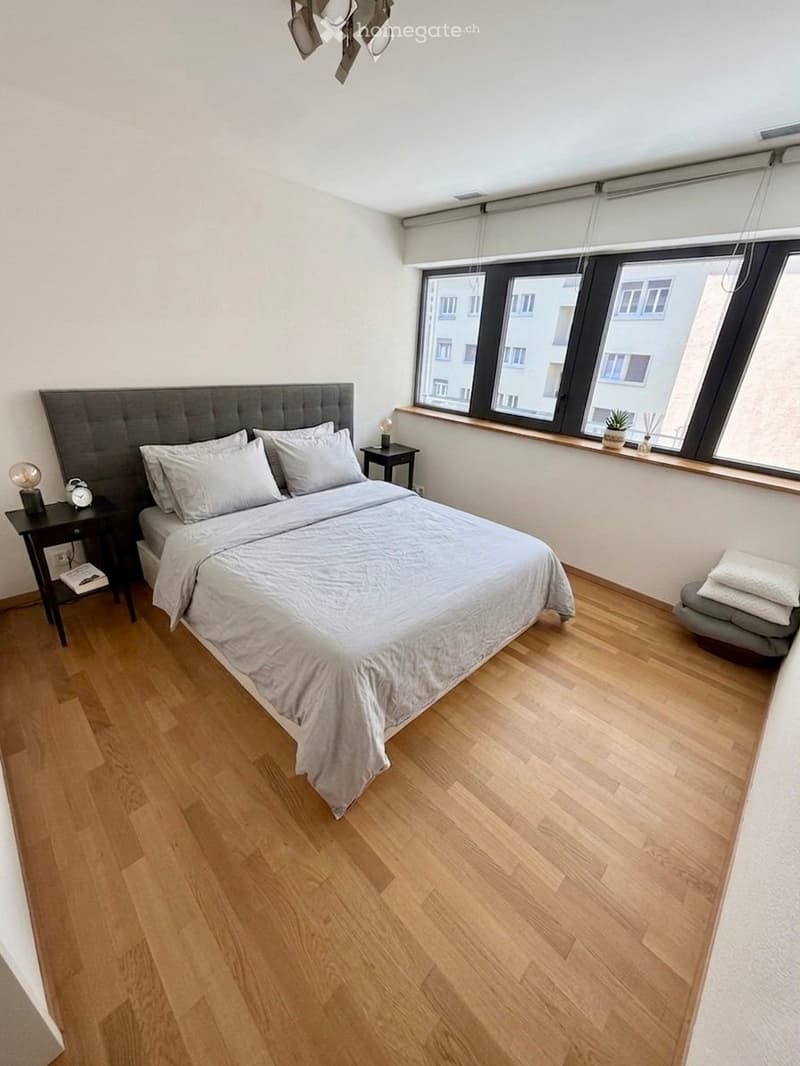 Modern bedroom with wooden flooring, large bed with grey headboard, nightstands, lamps, and a potted plant on the windowsill.