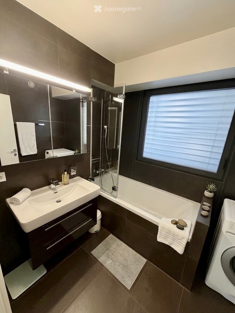 Modern bathroom with dark tiles, a large window, a bathtub, a sink, a mirror, and towels neatly placed on the floor and cabinet.